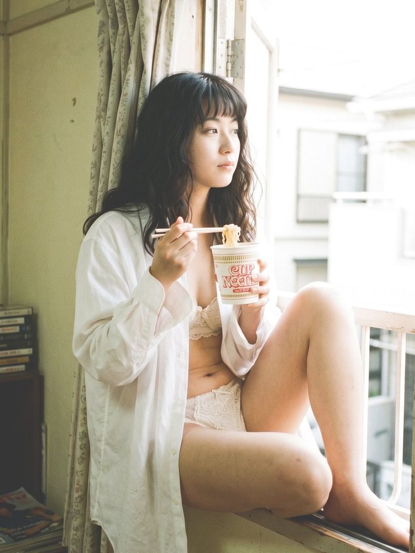 A young East Asian woman with long dark hair sits by a sunlit window wearing an open white shirt over white lace lingerie while eating cup noodles.