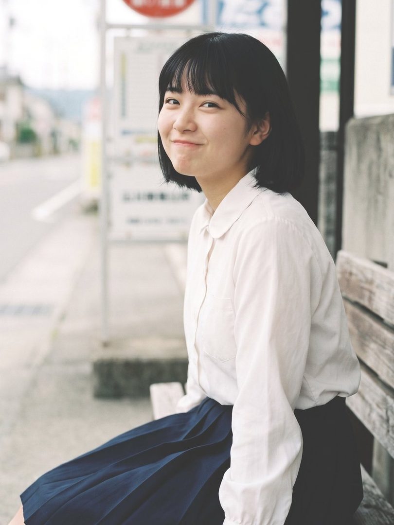 Young East Asian woman with short black bob hair wearing a white blouse and navy skirt, sitting on a wooden bench and smiling softly outdoors.