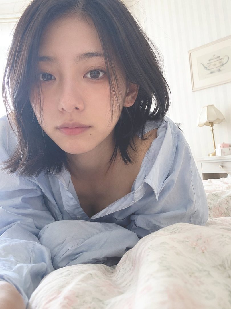 A young East Asian woman with short dark hair lies in bed wearing an open light blue shirt, captured in soft natural morning light.