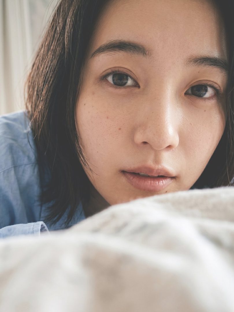 Close-up portrait of a young East Asian woman with freckles and dark hair, lying down and gazing at the camera in soft natural light.