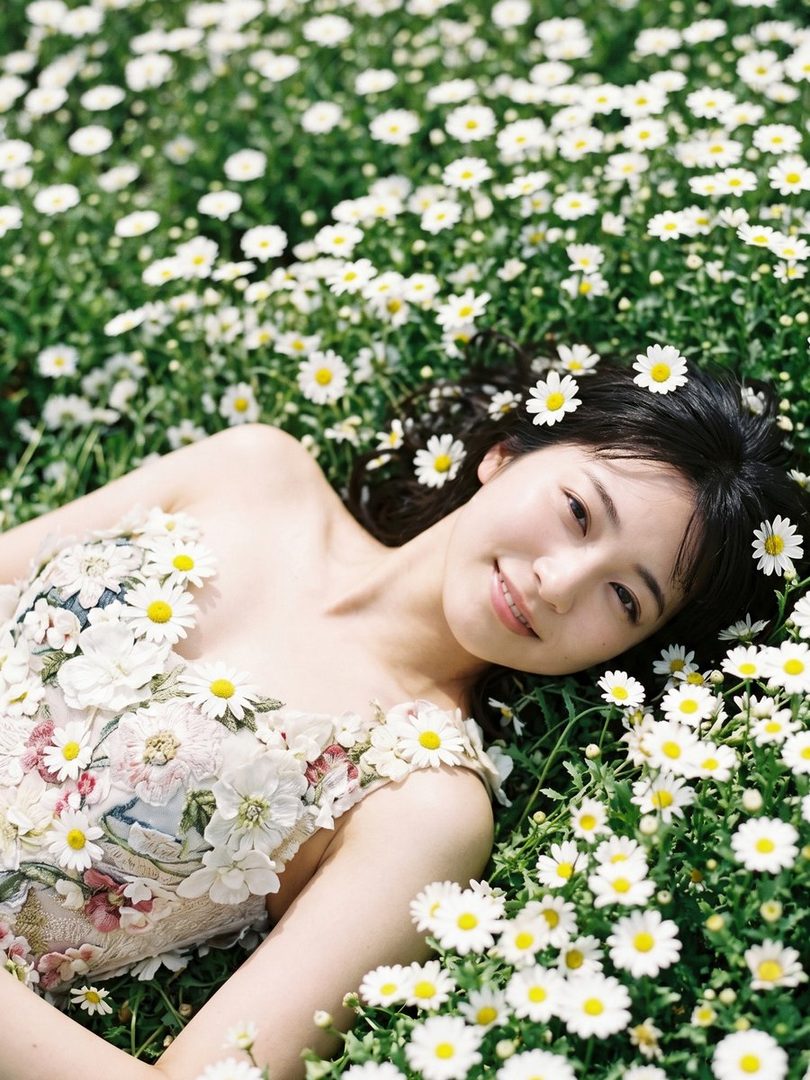 A young East Asian woman with dark wavy hair lies smiling in a lush field of white daisies, wearing a delicate floral-embroidered dress.