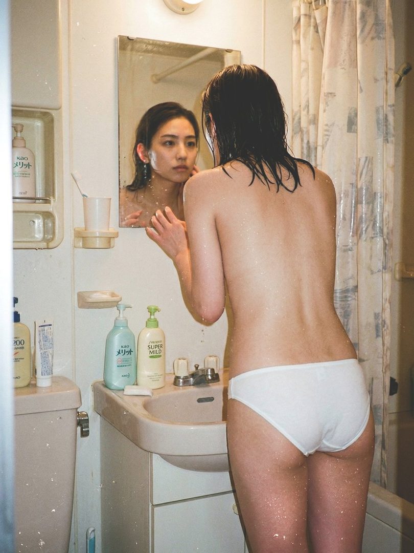 A young Asian woman with wet black hair stands topless in white underwear in a vintage bathroom, gazing at her reflection in the mirror.