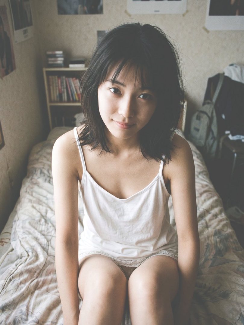 Young East Asian woman with short black hair in a white camisole, sitting on a bed in a cozy bedroom with soft natural light.