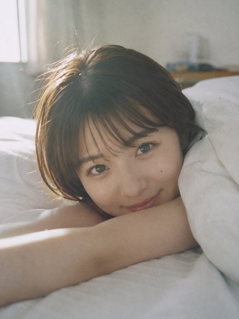A young East Asian woman with short brown hair rests her head on a white pillow in bed, smiling softly at the camera in soft morning light.