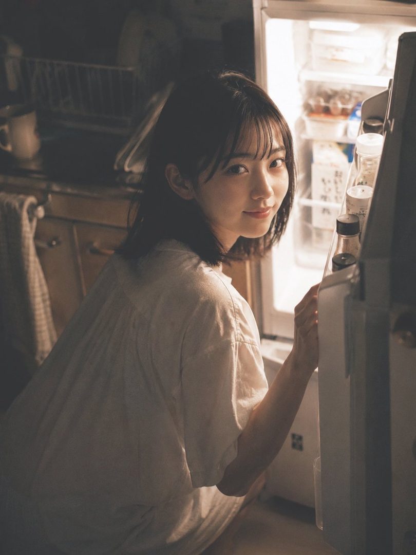 A young East Asian woman with short dark hair kneels in a dimly lit kitchen, smiling softly while illuminated by the light of an open refrigerator.
