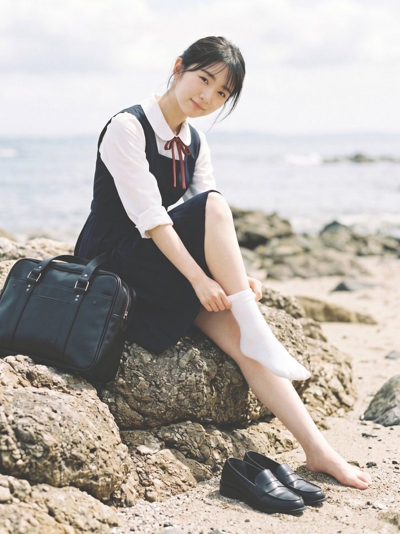 Young Japanese woman in vintage school uniform sitting barefoot on seaside rocks with a black bag nearby.