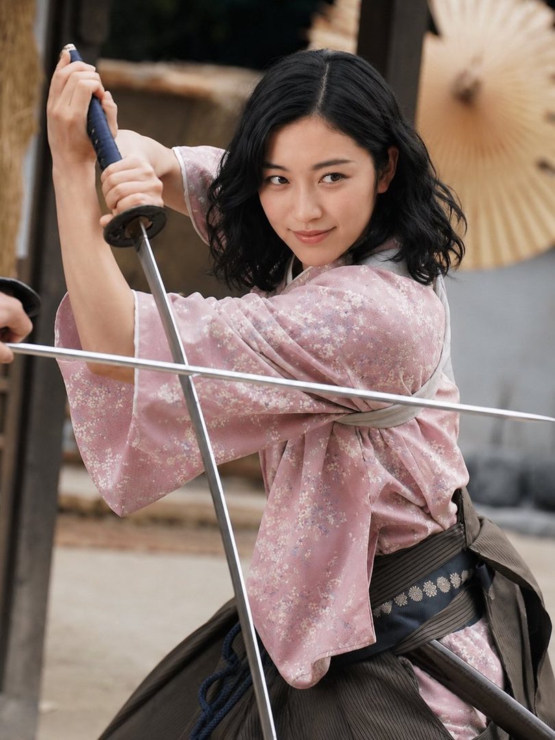 A young Japanese woman in a pink floral kimono and hakama poses dynamically with a drawn katana in a traditional Japanese setting.