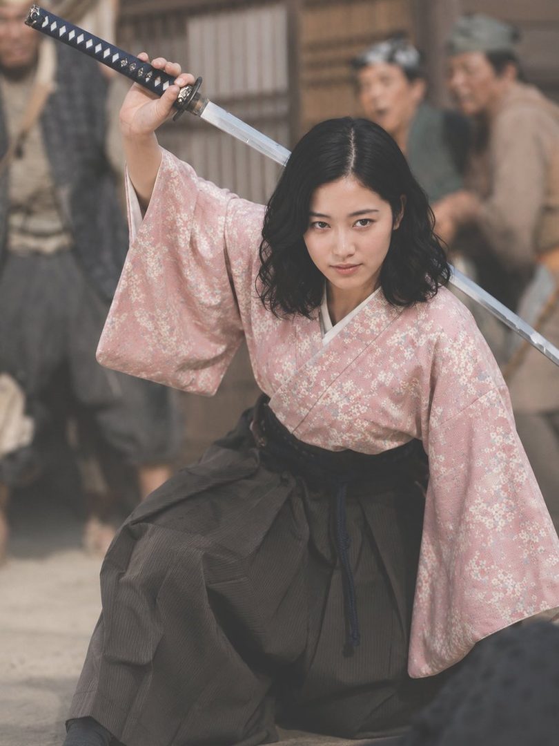 A young Japanese woman in a pink floral kimono raises a katana while kneeling in a historical street setting.
