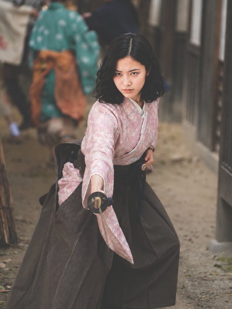 A determined young Japanese woman in pink floral kimono and hakama draws her katana on a historic street.