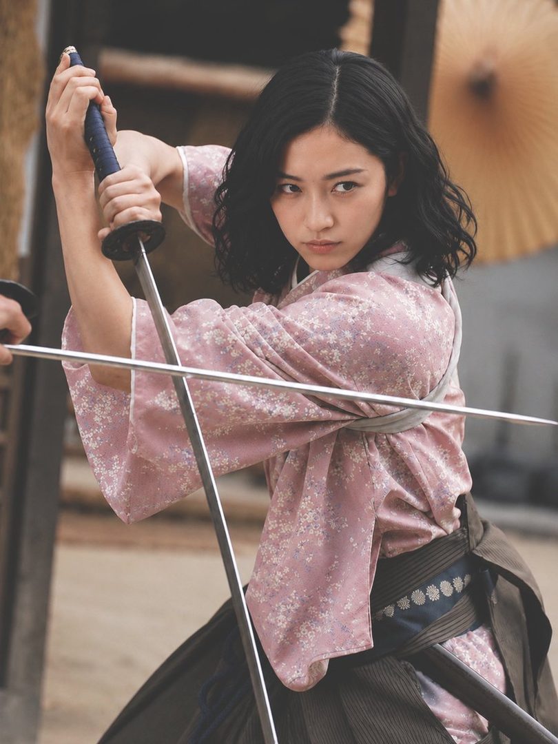 A young Japanese woman in pink floral samurai attire holding a katana in a dynamic fighting pose.
