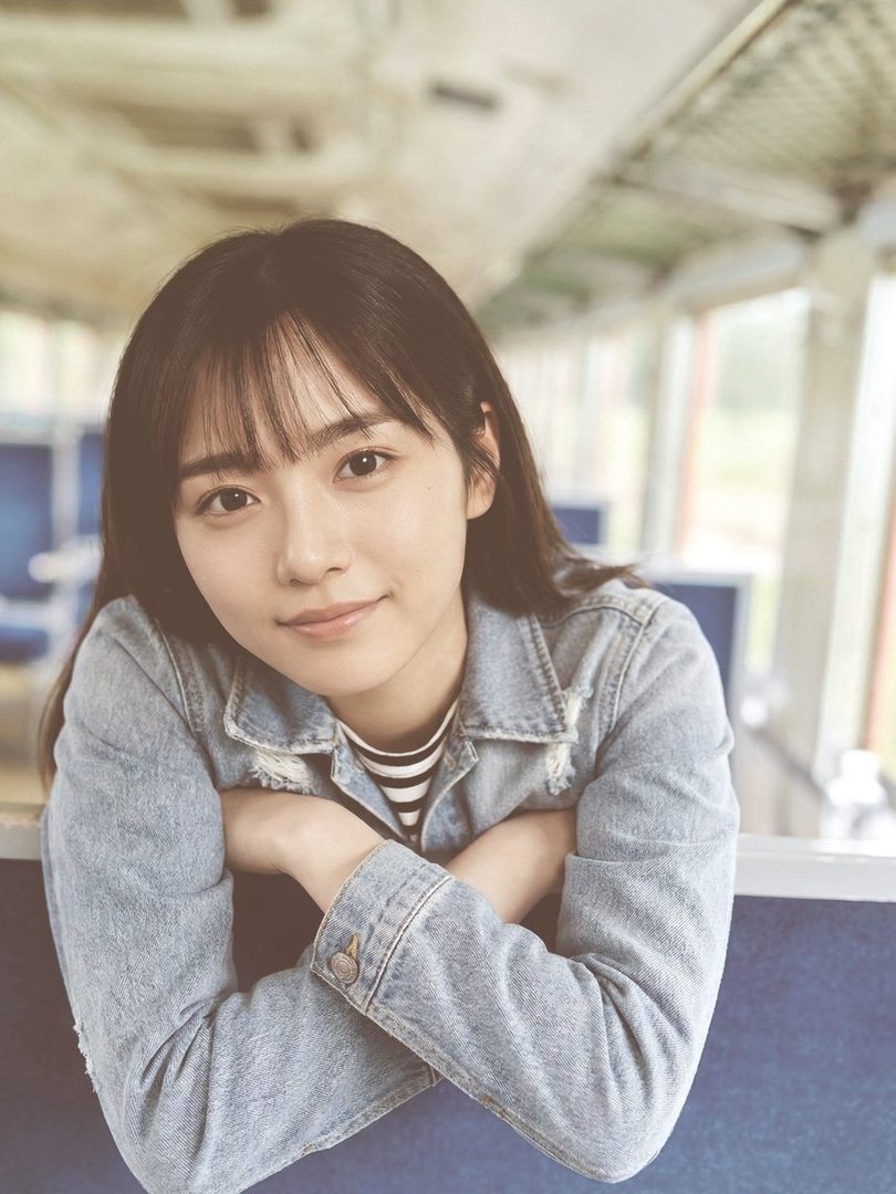 A young East Asian woman with dark shoulder-length hair and bangs wears a light denim jacket and smiles softly while leaning on a surface inside a train.