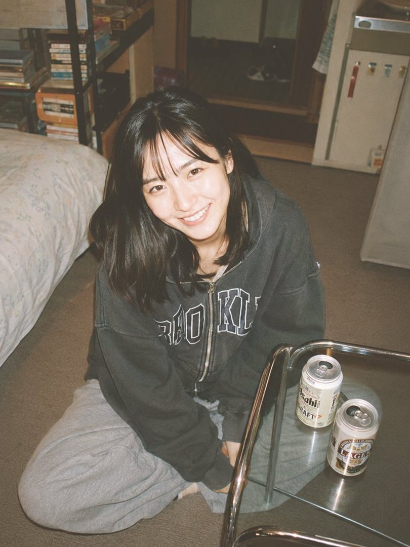 Young East Asian woman with shoulder-length black hair smiling while sitting casually on the floor in a cozy room with beer cans nearby.