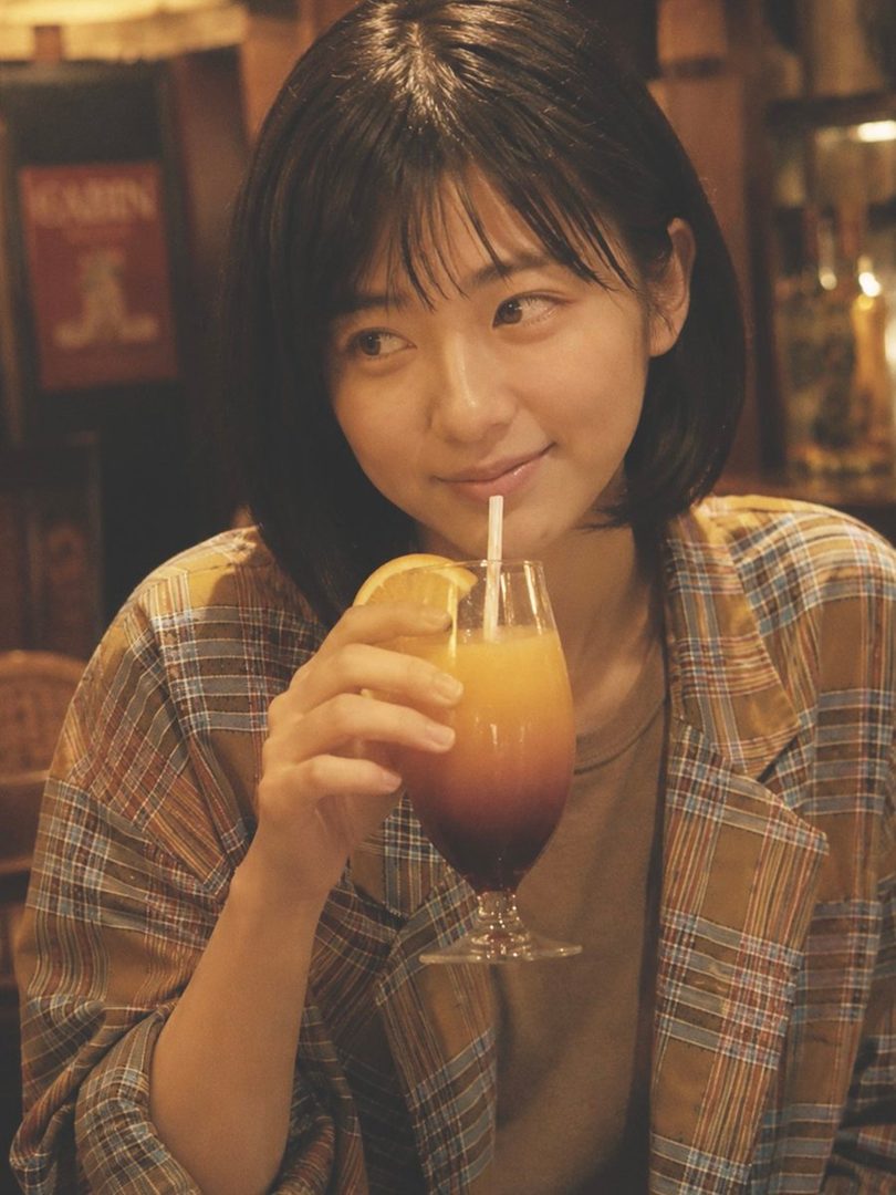 A young East Asian woman with short black hair smiles softly while holding a colorful sunset cocktail in a cozy retro bar.