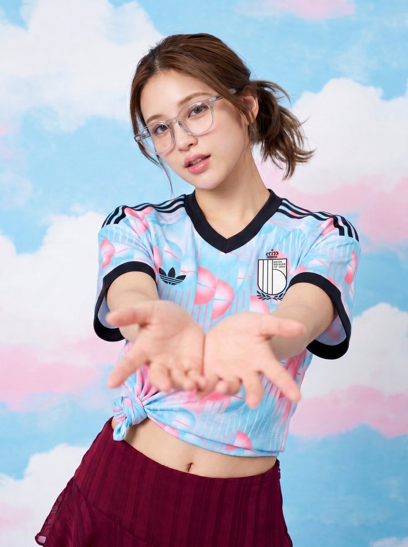 A young woman with brown hair in a ponytail and clear glasses wears a light blue and pink cropped t-shirt and a dark red skirt, posing against a blue and pink cloud-patterned background with her hands extended.