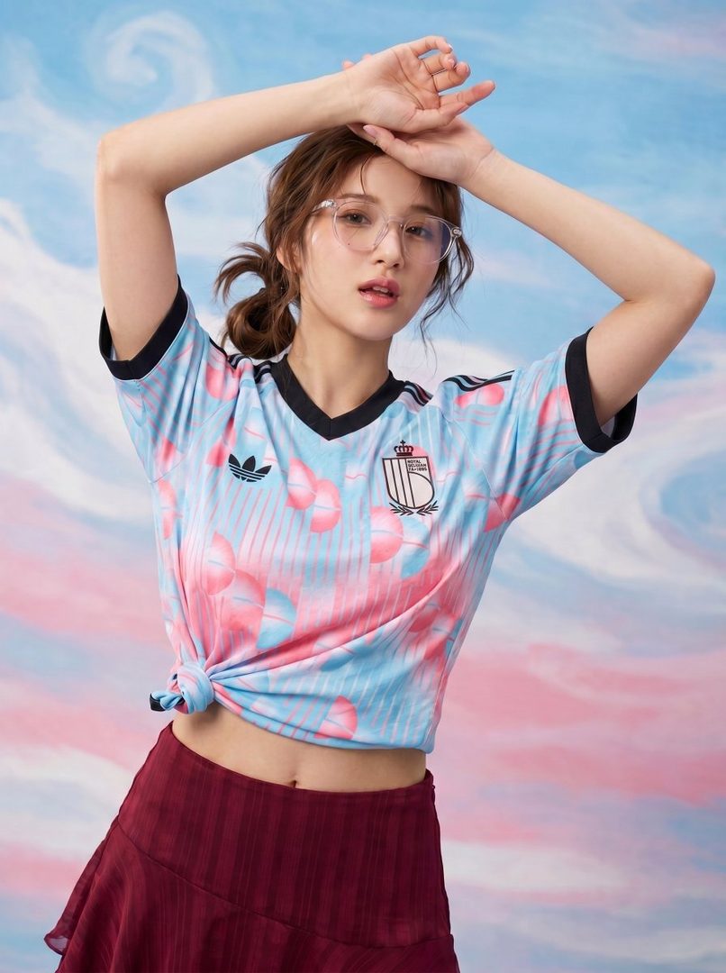 A young woman with brown hair in a messy bun and clear glasses poses in a light blue and pink cropped t-shirt and a maroon mini skirt against a pastel pink and blue background.