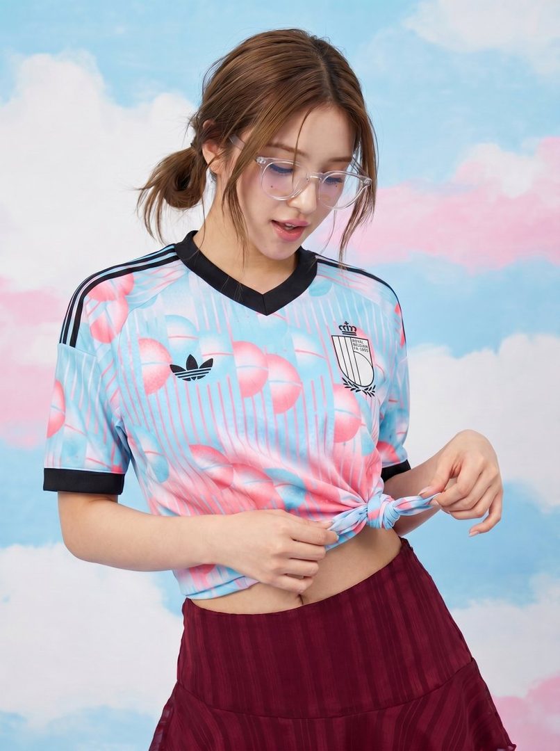 A young woman with brown hair in pigtails and clear glasses wears a light blue and pink patterned cropped t-shirt and a maroon mini skirt, posing against a pastel cloud background.