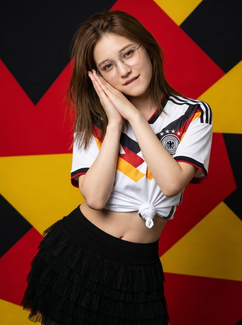 A young woman wearing a Germany FIFA World Cup jersey and a black tiered skirt, posing with her hands clasped under her chin against a black, red, and yellow geometric background.