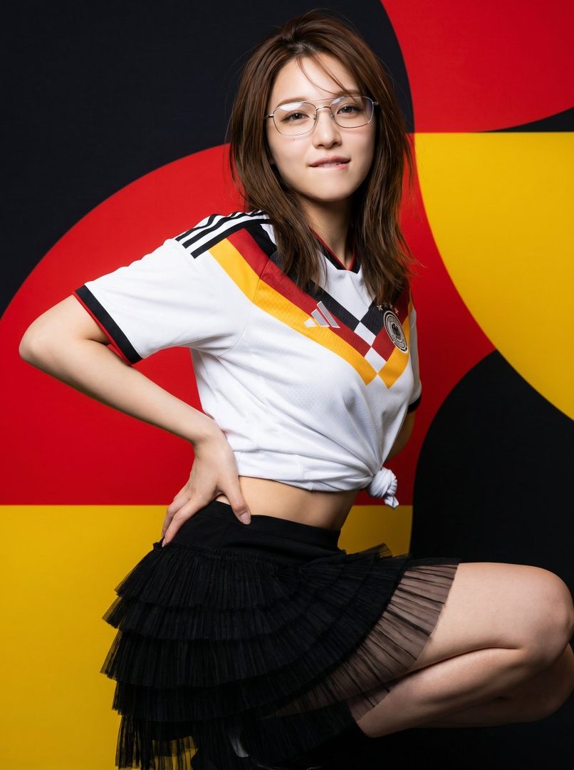 A woman wearing a white Germany FIFA World Cup jersey and a black tiered skirt, posing against a red, black, and yellow geometric background.