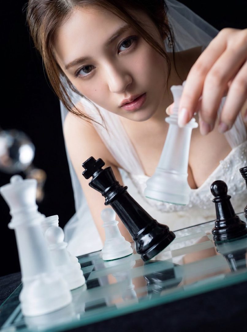 A woman in a white wedding dress plays chess, holding a clear queen piece over a black king piece on a transparent chessboard.