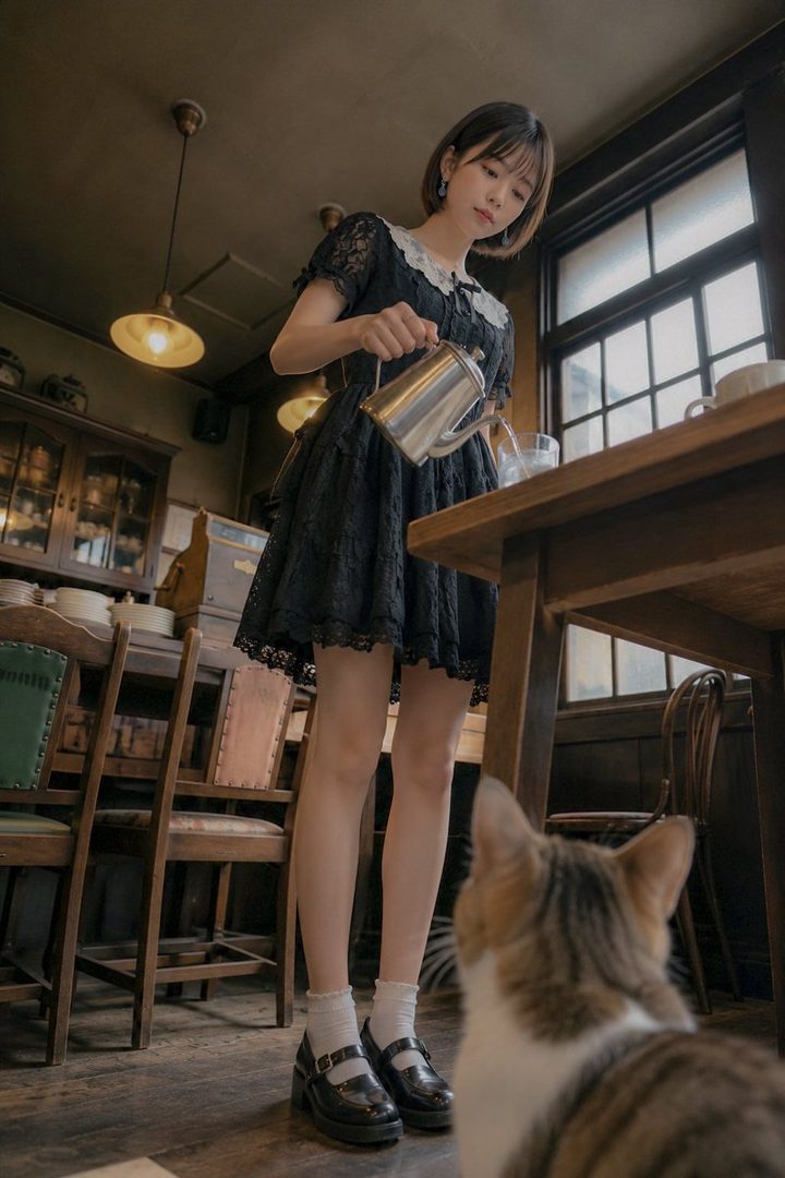 Young woman in a black lace dress pouring a beverage in a retro cafe while a cat watches from the foreground.