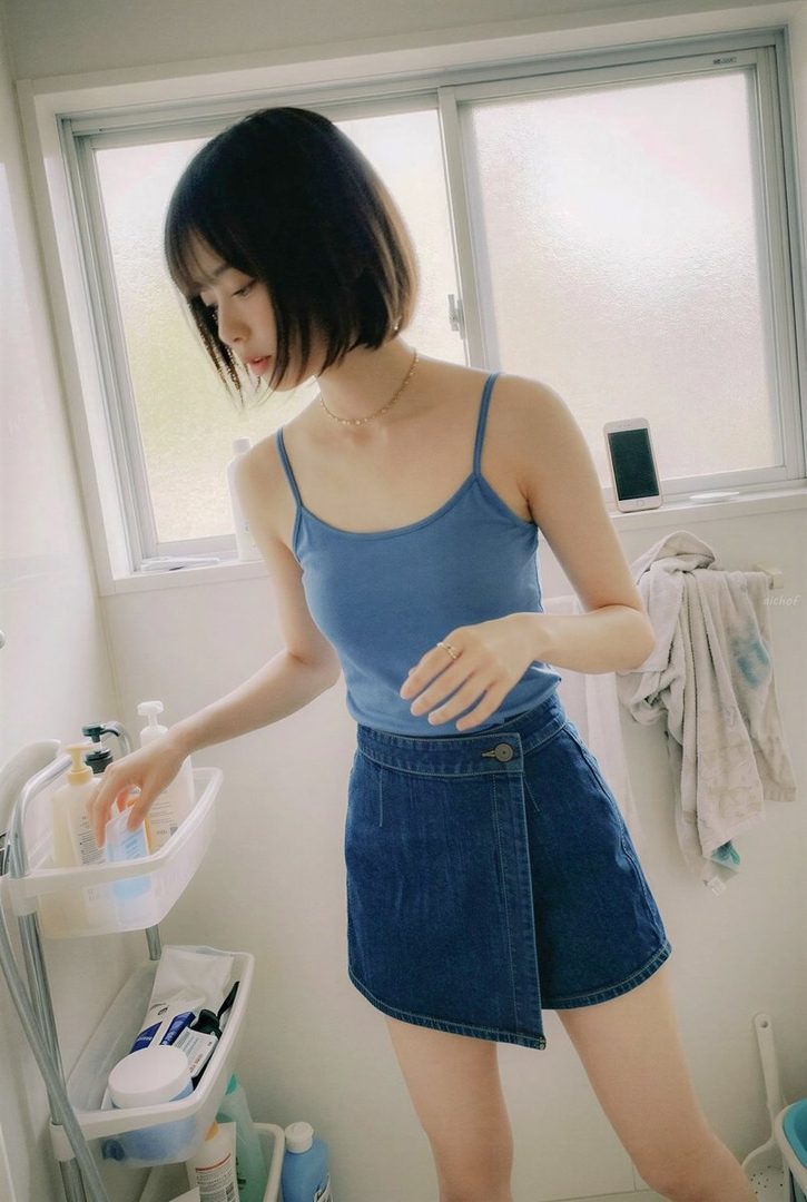 Young woman with short black bob hair wearing a blue camisole and denim wrap skirt in a bright bathroom.