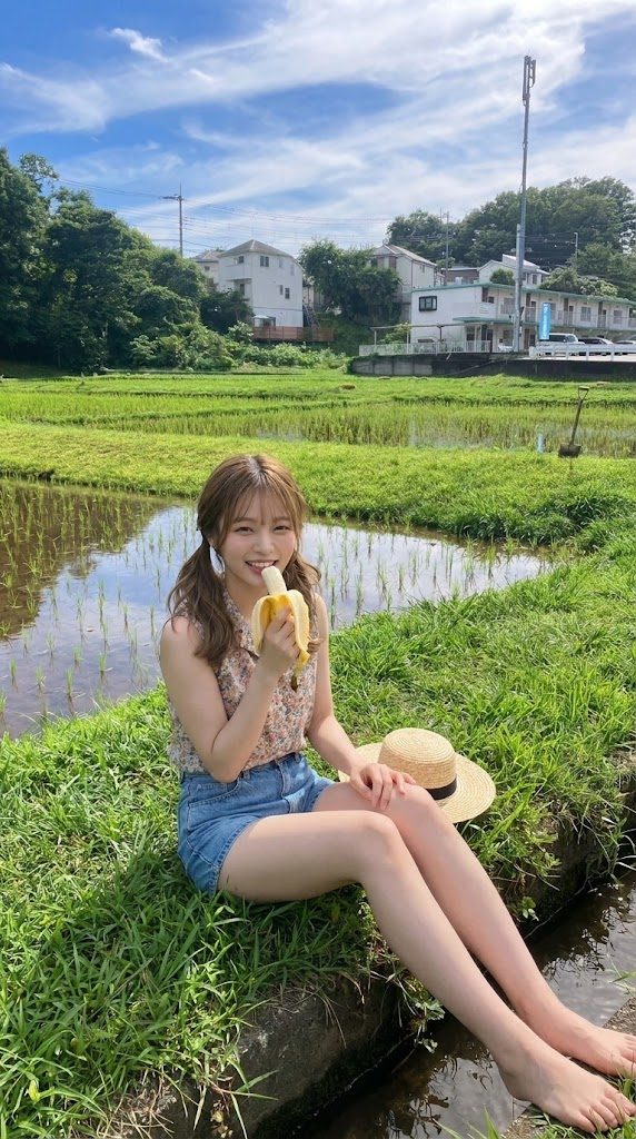 Young woman sitting barefoot by a rice paddy eating a banana on a sunny summer day in the countryside.