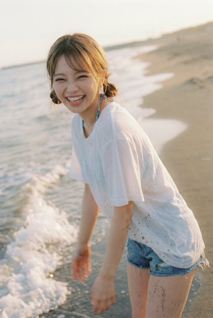 Smiling young woman with light brown pigtails in a wet white t-shirt and denim shorts standing playfully at the ocean's edge on a sunny beach.