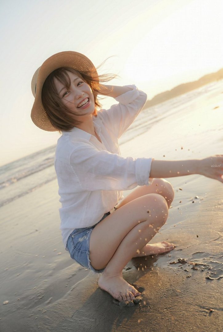 Smiling young woman in a straw hat and white shirt squatting barefoot on a sandy beach at golden hour sunset.