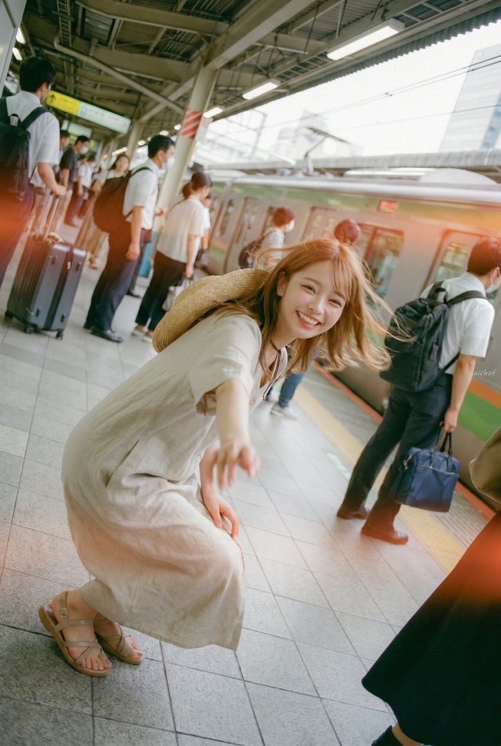 Young woman in a light dress smiling and reaching toward the camera on a busy Japanese train platform at sunset.