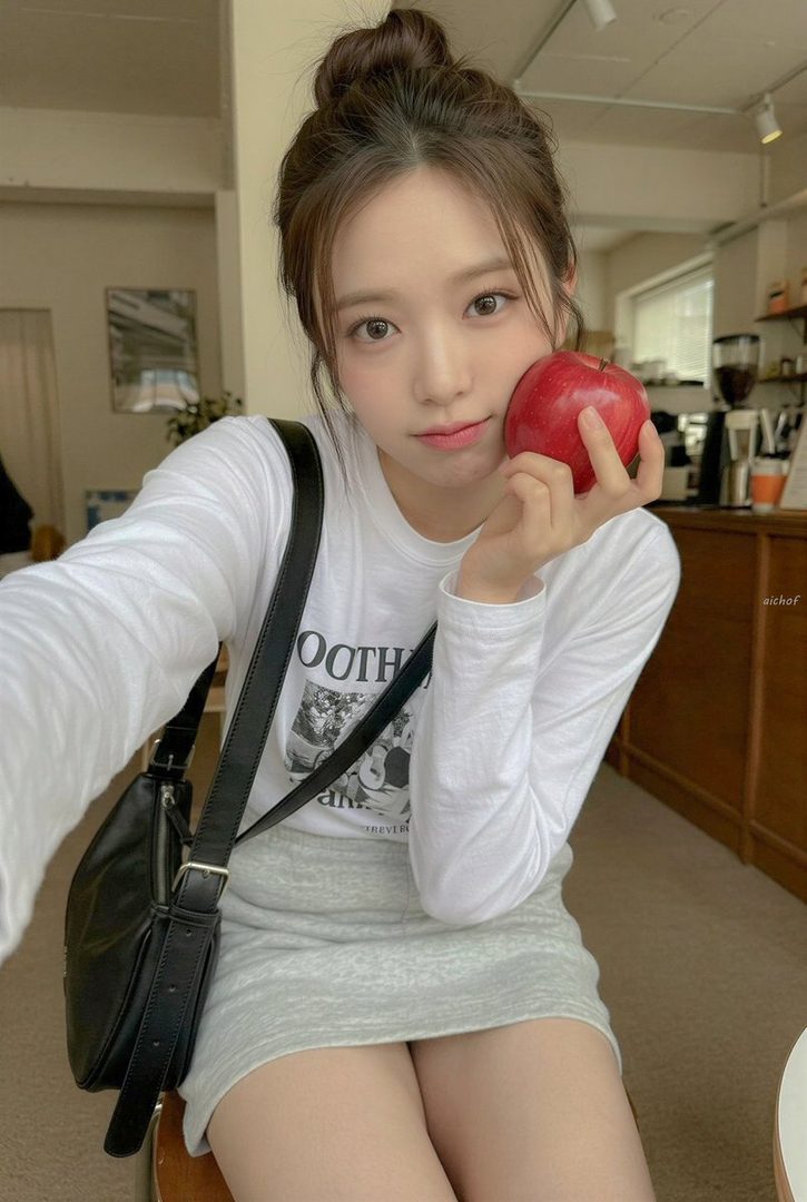 Young East Asian woman taking a casual selfie in a cafe while holding a shiny red apple against her cheek.