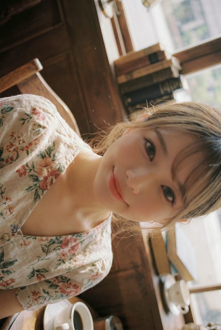 Young East Asian woman with light brown wavy hair and floral blouse smiling softly in a sunlit wooden cafe interior.