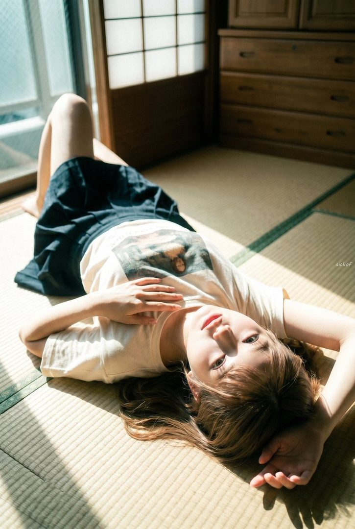 Young woman with long brown hair lying relaxed on a tatami mat in a sunlit Japanese room wearing a white t-shirt and dark skirt.