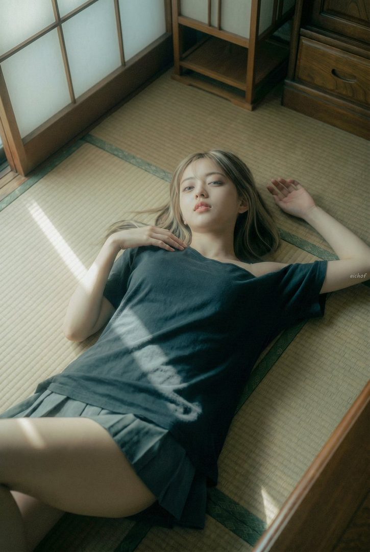 Young woman with long light brown hair lying relaxed on tatami in a sunlit traditional Japanese room, wearing a dark oversized t-shirt and shorts.