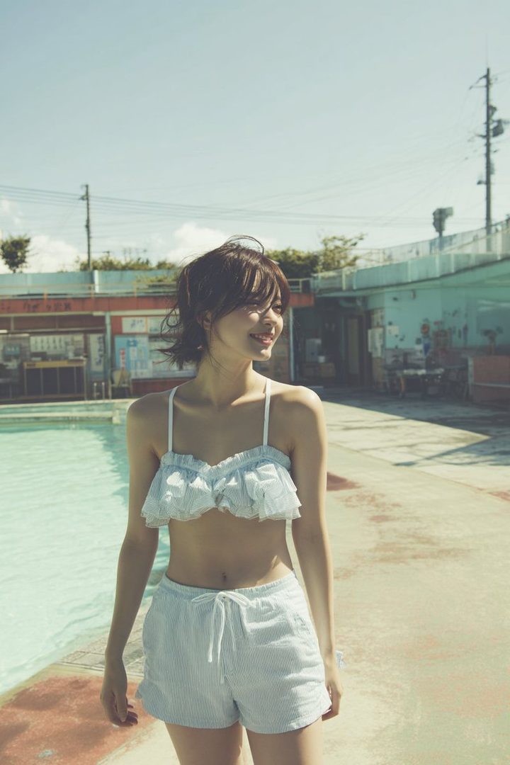 A young woman in a light blue frilled bikini top and striped shorts smiling by an outdoor pool on a sunny day.