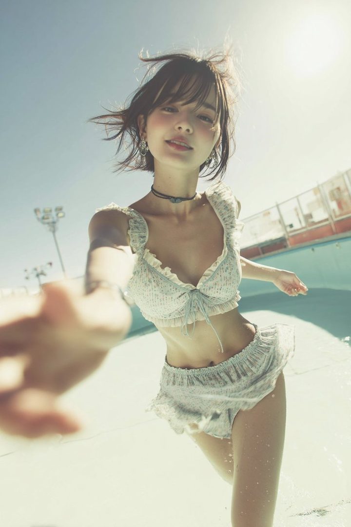 Young woman in a frilly white bikini taking a sunny selfie at an outdoor pool with water splashing around her.