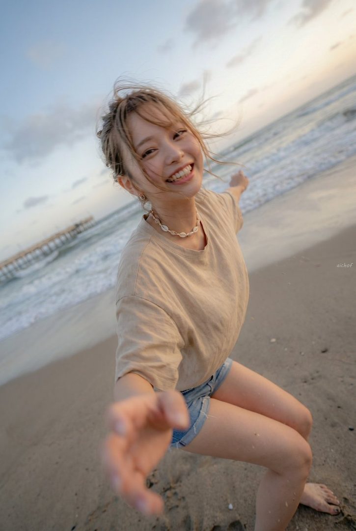Smiling young woman with wind-swept hair reaches toward the camera on a sunset beach.