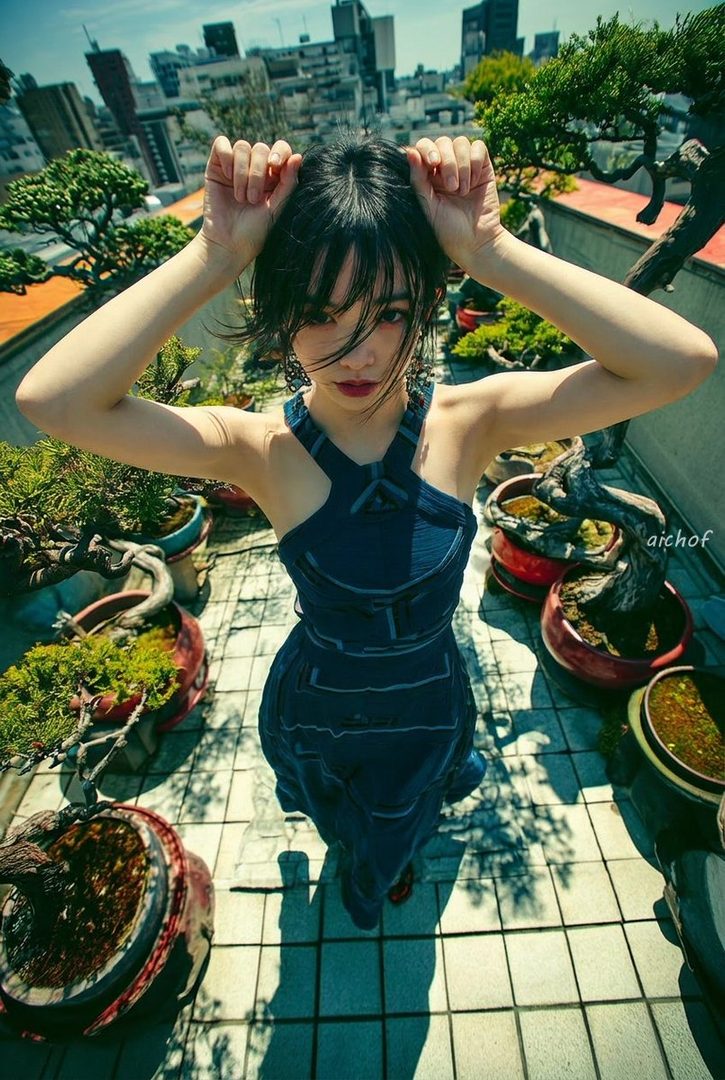 Young woman in a blue dress posing with hands in her hair on a sunny rooftop filled with bonsai trees overlooking a city.