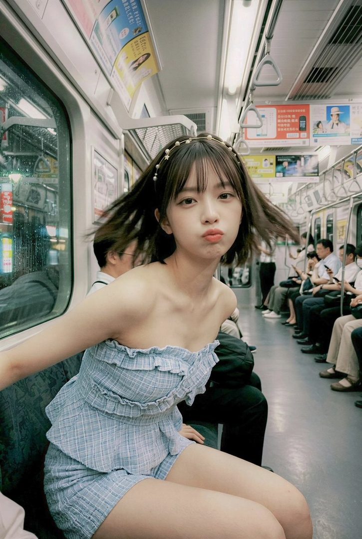 Young woman with short brown hair and pearl headband making a pouty face while seated in a Japanese commuter train wearing a blue gingham strapless outfit.
