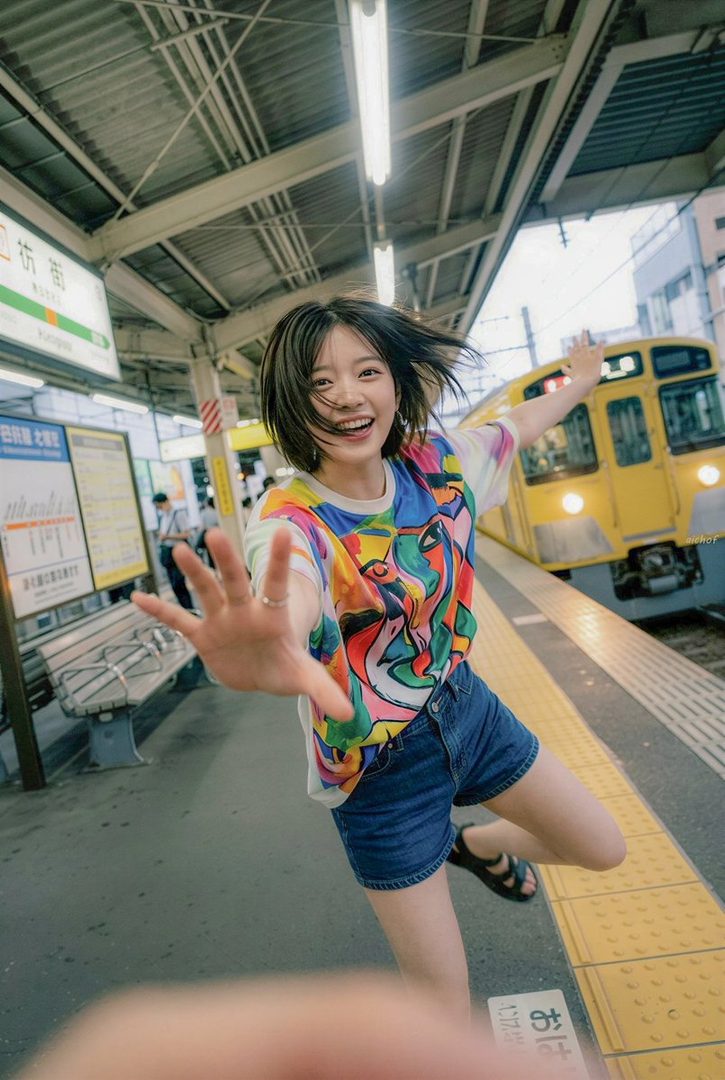 Young woman with short black hair joyfully reaching toward the camera on a sunny Japanese train platform beside a yellow train.