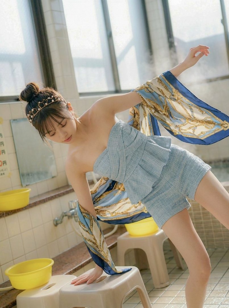 Young woman in light blue wrapped outfit striking a graceful pose inside a bright Japanese bathhouse with yellow basins.