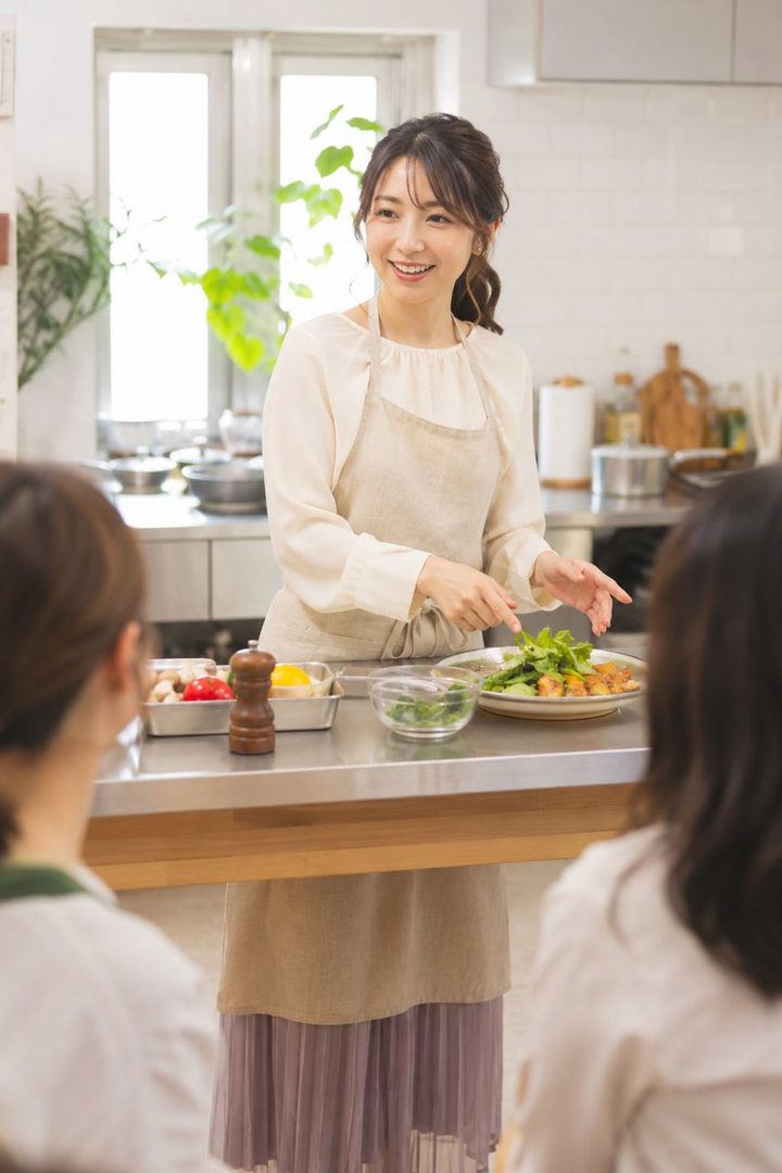 A smiling Japanese woman in her late twenties teaching a cooking class in a bright modern kitchen while wearing an apron.