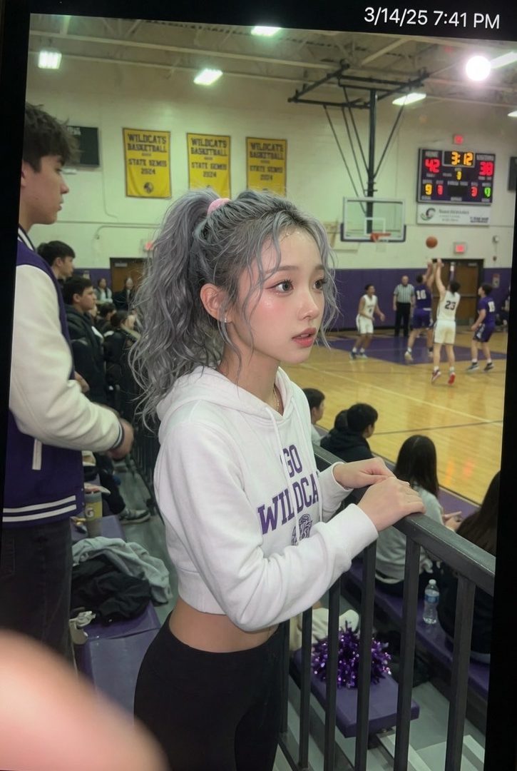 A young woman with silver hair in a ponytail, wearing a white cropped hoodie and black leggings, watches a basketball game from the stands.
