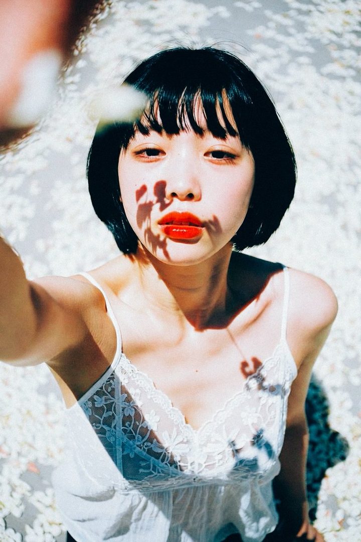 A young woman with short black hair and bright red lipstick takes a selfie outdoors, with a background of white flowers and dappled sunlight.