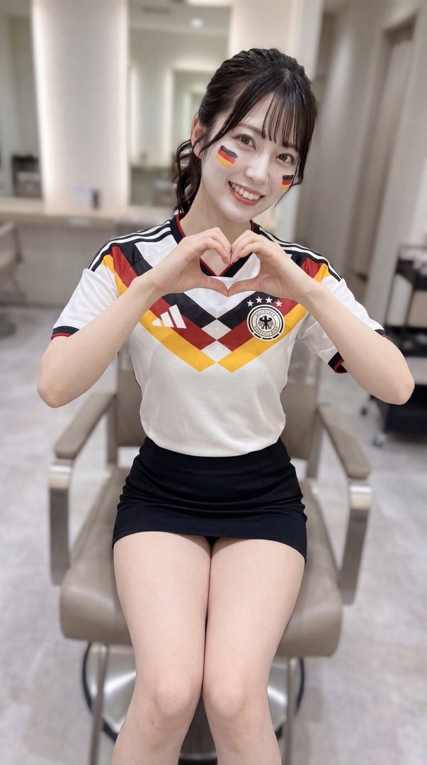 A young woman with German flag face paint smiles and makes a heart shape with her hands while wearing a German soccer jersey and a short black skirt.