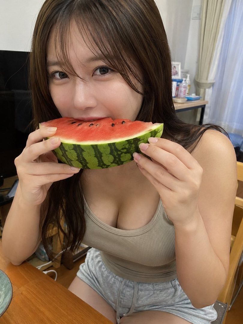 A young woman with long brown hair is eating a slice of watermelon indoors.