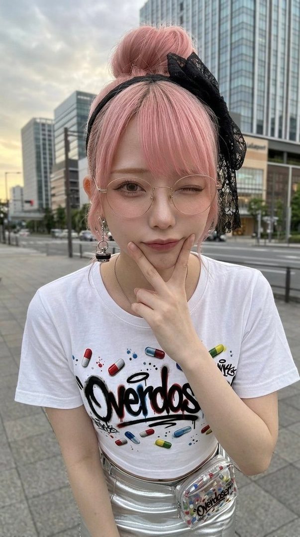 A young woman with pink hair styled in a bun, wearing glasses and a white t-shirt with a graphic print, poses playfully on a city street.