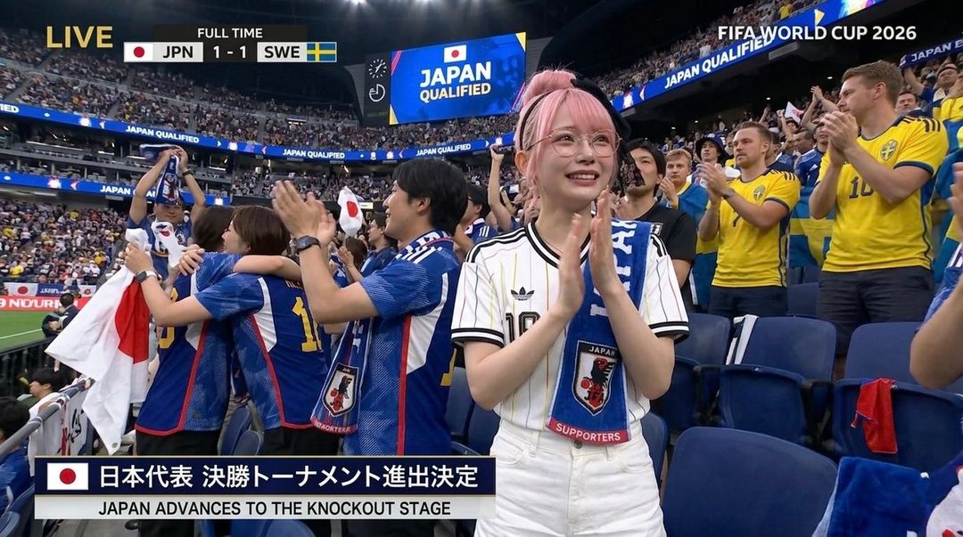 A young woman with pink hair and glasses smiles and claps in a stadium, wearing a Japanese soccer jersey and scarf.