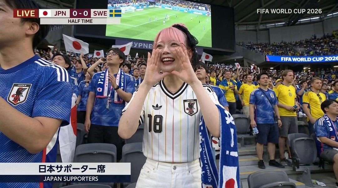 A young woman with pink hair and glasses cheers at a soccer match, wearing a Japanese jersey.