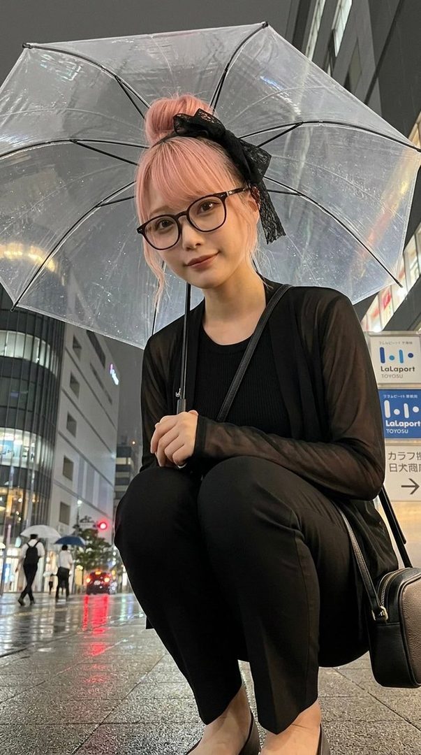 A young woman with pink hair tied in a bun, wearing glasses and a black outfit, crouches under a clear umbrella on a rainy city street at night.