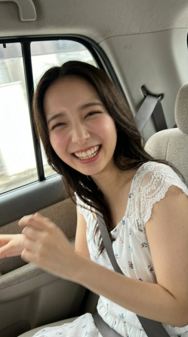 A young woman with long brown hair smiles broadly while sitting in the passenger seat of a car.